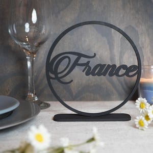 Op de afbeelding: Zwarte houten tafeldecoratie met het woord "France" uitgesneden in een cirkelvormig frame. Een brandende kaars, een wijnglas en een bord zijn ook zichtbaar. Madeliefjes zijn verspreid over de basis.