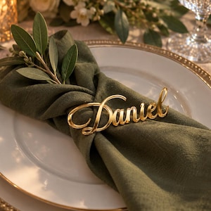 Può includere: Un segnaposto in acrilico color oro con il nome Daniel scritto in corsivo, appoggiato su un tovagliolo di stoffa verde oliva scuro piegato sopra un piatto piano bianco con bordo dorato e un piccolo rametto di foglie verdi.