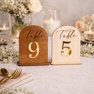 Può includere: Segnaposto numerici in legno con numeri dorati 9 e 5. I segnaposto sono arcuati nella parte superiore e presentano la parola "Table" in corsivo. Lo sfondo include fiori, candele e posate dorate.