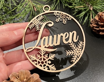 May include: A wooden Christmas ornament with the name "Lauren" cut out in a cursive font. The ornament is circular, with snowflake and pine branch designs around the edge. A small hole is at the top for hanging.