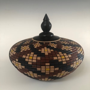 May include: A wooden box with a geometric pattern in shades of brown, black, and yellow. The box has a black wooden lid with a pointed top.