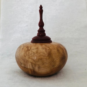 May include: A wooden urn with a cherry wood body and a red wood finial. The urn has a natural wood finish and a smooth, rounded shape.
