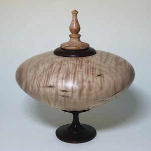 May include: A wooden lidded box with a light brown finish and a dark brown base. The box has a unique shape and a decorative finial on the lid.