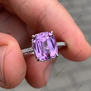 May include: A silver ring featuring a large, rectangular, light purple gemstone. The ring's band is adorned with small, clear stones. The ring is held up by a person's fingers.