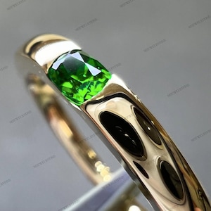Può includere: Un anello d'oro con una gemma verde brillante. La gemma, di forma rettangolare e con molteplici sfaccettature, è incastonata in una fascia d'oro lucida. Il design dell'anello è semplice ed elegante, che mette in risalto la pietra verde.