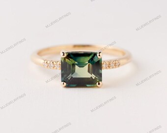May include: A gold ring with a square-cut, dark green gemstone. The band is embellished with small, clear accent stones on either side of the main stone. The ring is set against a white backdrop, accentuating the jewellery's design.