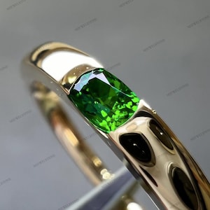 May include: A gold ring with a bright green gemstone. The ring's band is polished gold, reflecting light. The gemstone is rectangular and faceted, displaying its vivid green hue. The ring is a piece of jewellery.