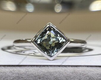 May include: A silver ring with a square-cut, pale green gemstone. The gemstone is set in a square bezel, and the band is polished silver. The ring is shown on a white surface.