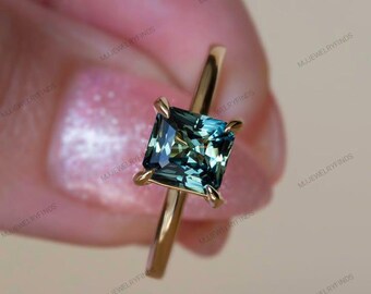 May include: A gold ring with a square, teal gemstone. The stone is held in place by four prongs, and the band is plain gold. The ring is shown against a blurred background.