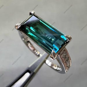 Pode incluir: Um anel de prata com uma grande pedra preciosa retangular de cor verde-azulado. A pedra preciosa tem uma qualidade vibrante e reflexiva. A banda do anel tem um design detalhado e texturizado com pequenas pedras de acento claras.