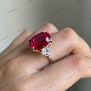 May include: A large, oval-cut ruby ring with a gold setting and a smaller, heart-shaped diamond accent. The ring's band is silver. The ruby is a deep red color, and the diamonds sparkle. The ring is a piece of fine jewelry.