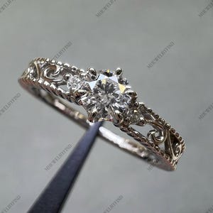 May include: A silver engagement ring with a large, round, clear gemstone at the centre, flanked by smaller stones. The band features intricate filigree and rope-like detailing, creating a vintage aesthetic.