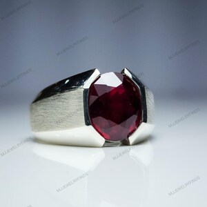 May include: A silver ring with a large, oval-cut, deep red gemstone. The ring has a brushed metal finish on the band and a polished surface around the gemstone. The gemstone is faceted, catching the light and creating a sparkling effect.
