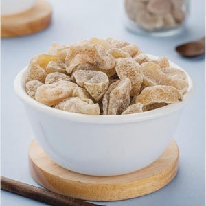 May include: A white ceramic bowl filled with candied ginger pieces. The ginger is light brown and has a crystallized sugar coating. The bowl sits on a small wooden coaster, with a light blue background.