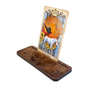 Puede incluir: Un soporte de madera para cartas del tarot con un diseño decorativo de polilla y sol. El soporte sostiene una carta del tarot con una ilustración del sol y el texto "THE SUN". La carta está inclinada, mostrando la obra de arte.