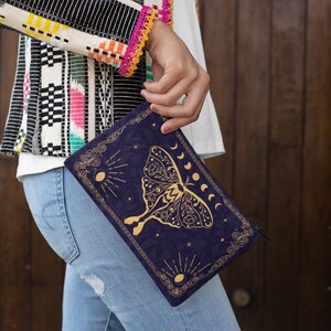 Pu&ograve; includere: Una pochette blu scuro con una cerniera dorata e un design dorato di una falena con fasi lunari e stelle. Il design &egrave; su uno sfondo blu scuro.