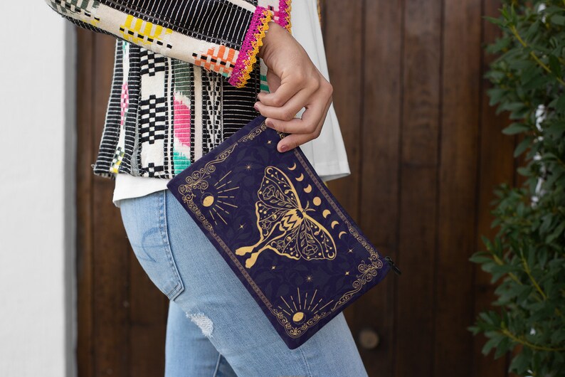 Pu&ograve; includere: Una pochette blu scuro con una cerniera dorata e un design dorato di una falena con fasi lunari e stelle. Il design &egrave; su uno sfondo blu scuro.
