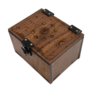 May include: A wooden box with a celestial design etched on the lid. The box is a dark brown color and features a sun, moon phases, and stars. The lid is secured with black leather straps and a metal clasp. The box is likely used for storing small items.