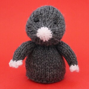 May include: Hand-knitted gray mole plush toy with a white pom-pom nose and white accents on the paws. The mole has black button eyes and is set against a solid red background. A cute, handmade stuffed animal.