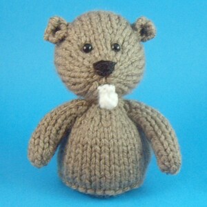 May include: A brown knitted stuffed animal resembling a beaver with black eyes, a black nose, and a white mouth.