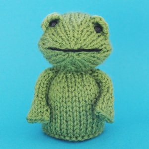 May include: A green knitted frog toy with black eyes. The frog is sitting on a blue background.