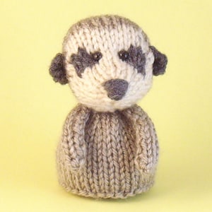 May include: A knitted meerkat toy with brown and white fur, black eyes, and a gray nose. The toy is sitting on a yellow background.