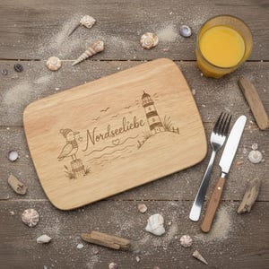 Könnte beinhalten: Ein rechteckiges Holzschneidebrett mit einer Lasergravur, die eine Möwe mit Mütze, einen Leuchtturm und den Schriftzug Nordseeliebe zeigt. Das Brett wird von einer Edelstahlgabel und einem Messer mit Holzgriff ergänzt.