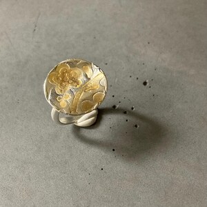 May include: A silver ring with a round, textured face featuring a gold floral design. The ring's band has a twisted, rope-like appearance. The ring is set against a gray background, casting a shadow.