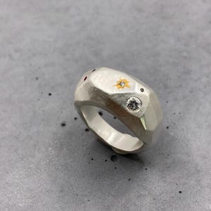 Puede incluir: Un anillo de plata con un diseño facetado, con una estrella dorada con un diamante central, un diamante transparente y dos pequeñas gemas rojas. El anillo tiene un acabado de metal cepillado y una banda ancha.