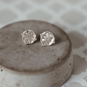 May include: A pair of silver-colored stud earrings. Each earring features a circular design with a floral or sunburst pattern in the center. The earrings are displayed on a gray, circular surface.