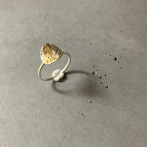 May include: A silver ring with a gold-colored, teardrop-shaped centerpiece. The gold section features an embossed floral design. The ring is set against a gray surface, with a small white object supporting the ring.