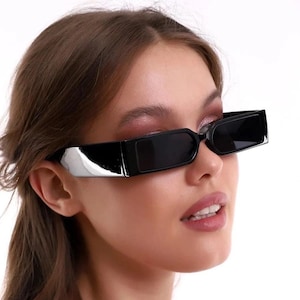 Peut inclure: Lunettes de soleil rectangulaires noires avec un accent argenté sur les branches. Les lunettes de soleil ont des verres foncés et une monture épaisse. L'image est un gros plan des lunettes de soleil.