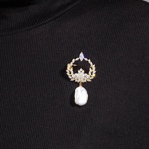Op de afbeelding: Goudkleurige broche met een lauwerkransontwerp, heldere en paarse accenten en een grote, witte, druppelvormige parel. De broche is vastgemaakt aan een zwarte coltrui.