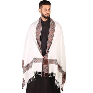 May include: White shawl with a brown, red, and blue patterned border. The shawl has a fringe on the bottom edge.