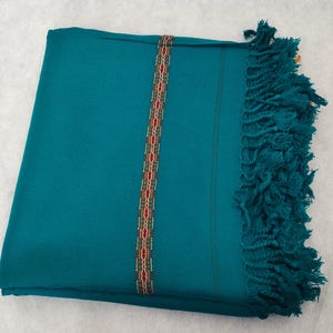 May include: A teal blue shawl with a red, gold, and white patterned border. The shawl has fringe on one side.