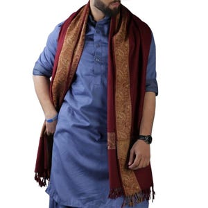 May include: A burgundy scarf with a gold paisley border and fringe. The scarf is draped over a blue shirt. The scarf is a warm accessory for cooler weather.