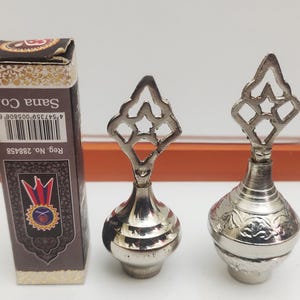 May include: Two silver metal perfume bottles with intricate designs. The bottles have a round body and a decorative top. The bottles are on a white surface.