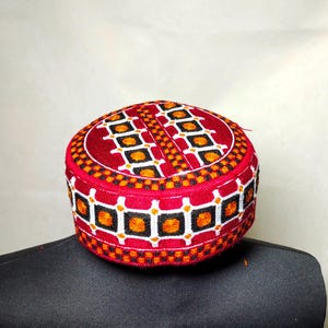 May include: A red, black, and white embroidered cap with a geometric pattern. The cap is made of a soft, textured fabric.
