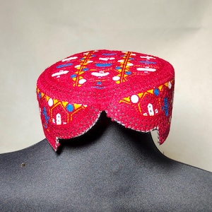Puede incluir: Un gorro de fieltro rojo con un diseño bordado en blanco, azul y amarillo. El gorro tiene un borde festoneado y se lleva sobre un maniquí negro.