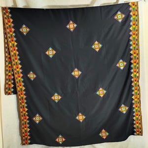 Puede incluir: Textil negro con patrones de diamantes bordados coloridos. Los bordes presentan un borde detallado con diseños geométricos en rojo, amarillo, verde y naranja. La tela está drapeada, mostrando el intrincado bordado.