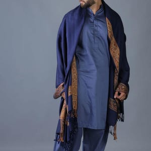 May include: A navy blue shawl with a gold and brown paisley border. The shawl is draped over a person wearing a blue shirt and pants.