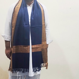 May include: A navy blue scarf with a gold border and fringe. The scarf is draped over a white shirt. The gold border has a detailed pattern. The scarf is a rectangular shape and appears to be made of a soft material.