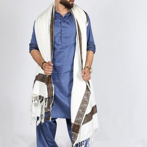May include: A white shawl with a brown paisley pattern border. The shawl is draped over a person wearing a blue shirt and pants.