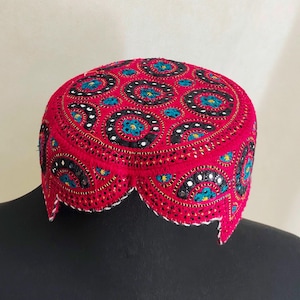 May include: A vibrant red embroidered cap with a circular pattern in black, white, and teal. The cap has a scalloped edge with white trim. The intricate design suggests a cultural or traditional style.
