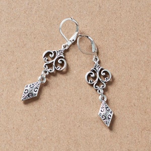 Puede incluir: Un par de pendientes de plata con un diseño delicado y ornamentado. Cada pendiente presenta un colgante en forma de diamante con un patrón de filigrana y un colgante más pequeño e intrincado por encima.