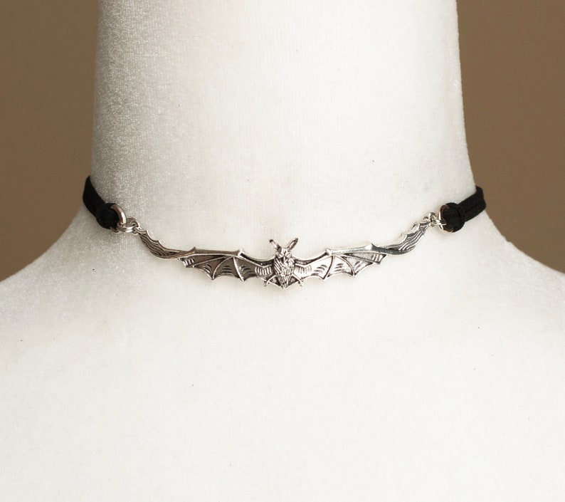 May include: A black cord choker necklace featuring a central silver-toned metal bat pendant with outstretched wings. The bat is detailed with textured wing patterns and is attached to the black cord via small silver-toned metal jump rings.