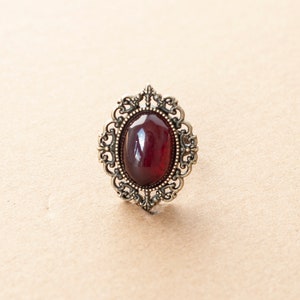 May include: A vintage-style ring with a dark red oval gemstone set in an ornate silver-toned filigree design.
