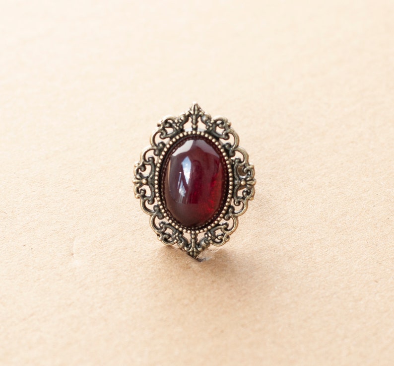 May include: A vintage-style ring with a dark red oval gemstone set in an ornate silver-toned filigree design.