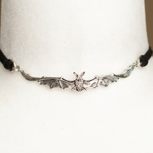 May include: A choker necklace featuring a silver-toned metal bat pendant with outstretched wings. The pendant is attached to a thin black cord on both sides, creating a horizontal design that rests against the neck with a detailed bat figure.
