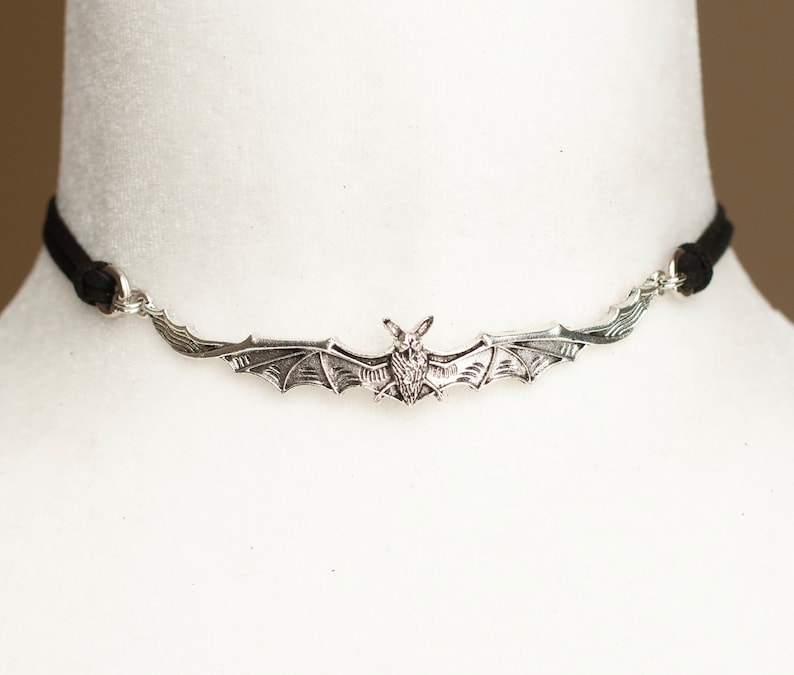 May include: A choker necklace featuring a silver-toned metal bat pendant with outstretched wings. The pendant is attached to a thin black cord on both sides, creating a horizontal design that rests against the neck with a detailed bat figure.